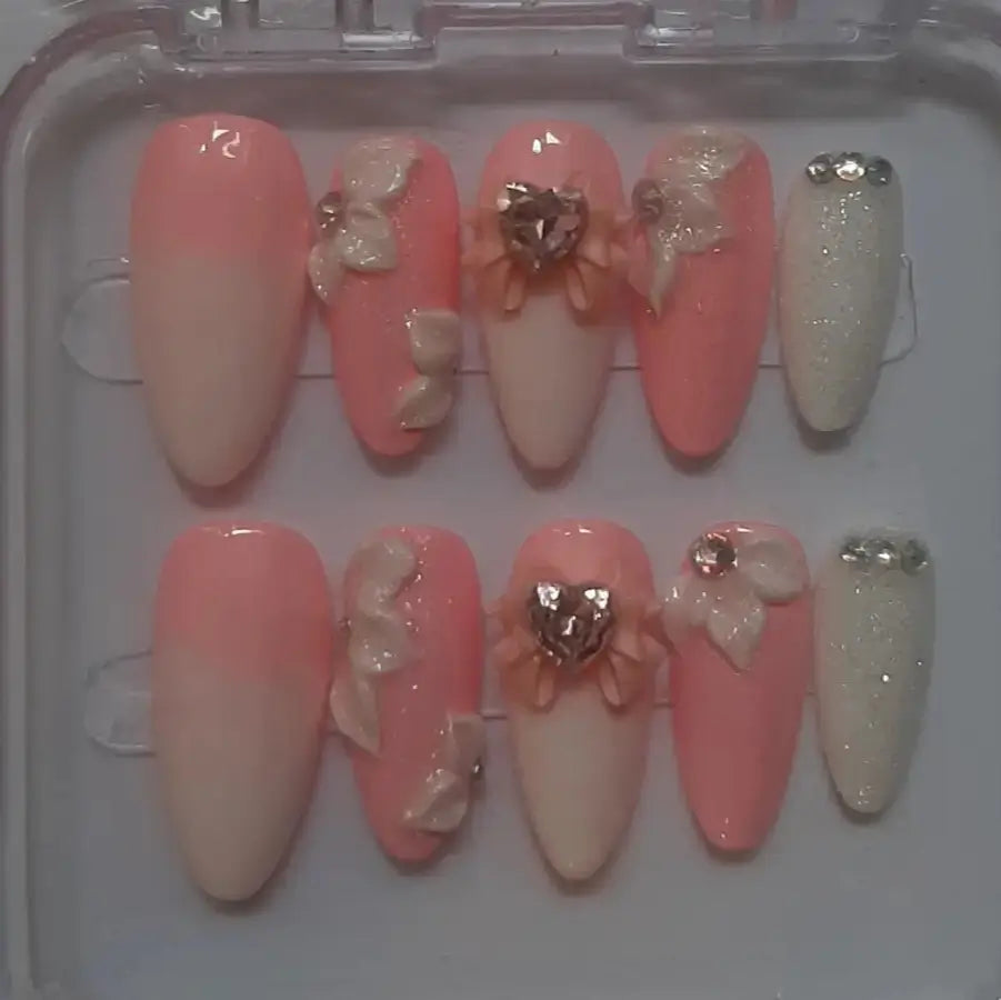 Set de 10 faux ongles Baby Pink dans leur boîtier transparent, présentant des designs romantiques en nuances de rose et nude avec strass, nœuds et ornements dorés, finition brillante professionnelle