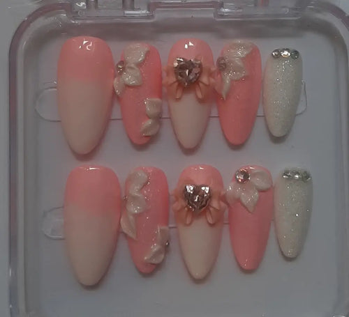 Set de 10 faux ongles Baby Pink dans leur boîtier transparent, présentant des designs romantiques en nuances de rose et nude avec strass, nœuds et ornements dorés, finition brillante professionnelle