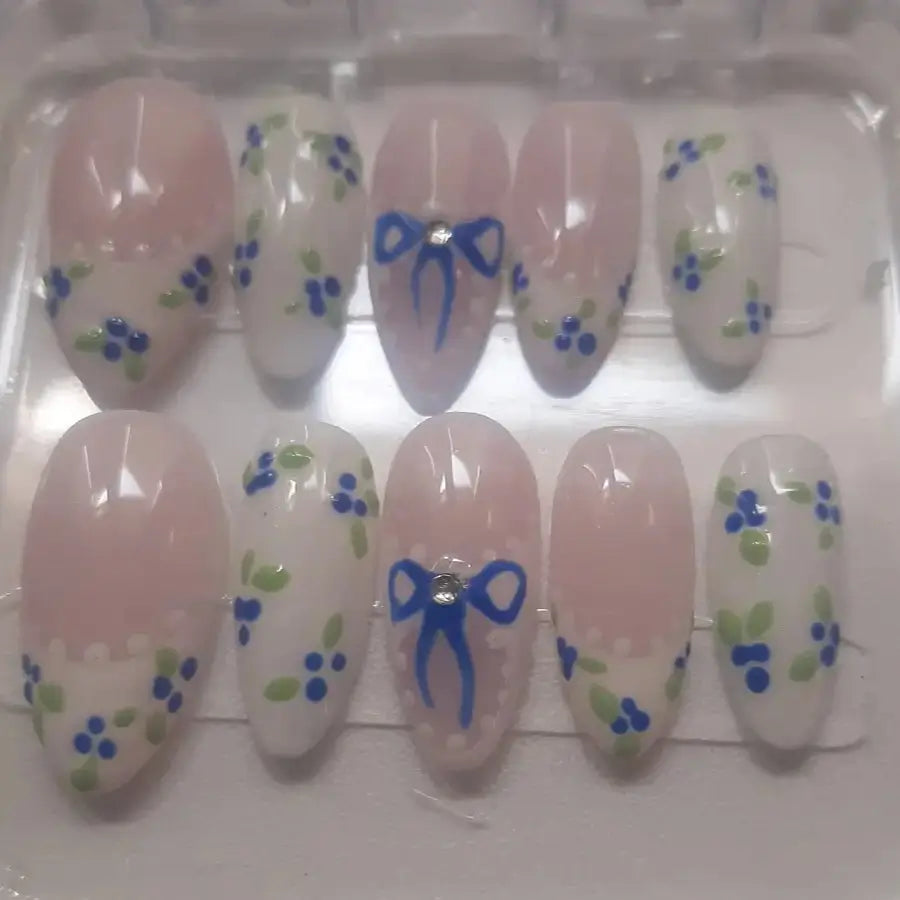 Craquez pour ces adorables faux ongles aux tons poudrés ornés de délicates myrtilles bleues et de nœuds charmants qui évoquent un jardin anglais en été.
