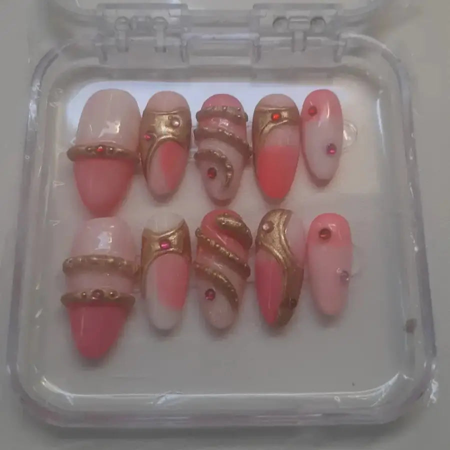 Set de 10 press-on nails présentant des ongles dans des tons roses nude et corail, ornés de motifs serpentins dorés métallisés et de strass rouges.