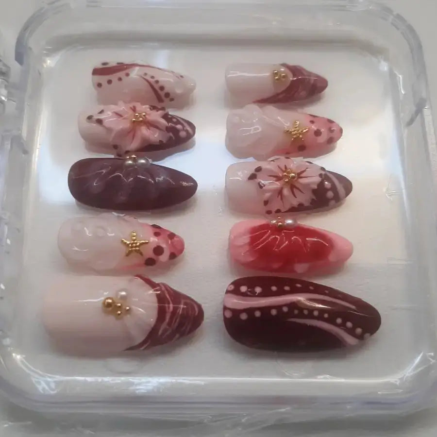 Set de 10 press-on nails dans un étui transparent, présentant des ongles dans des tons roses, bordeaux et nude, ornés de motifs floraux 3D avec pétales délicats
