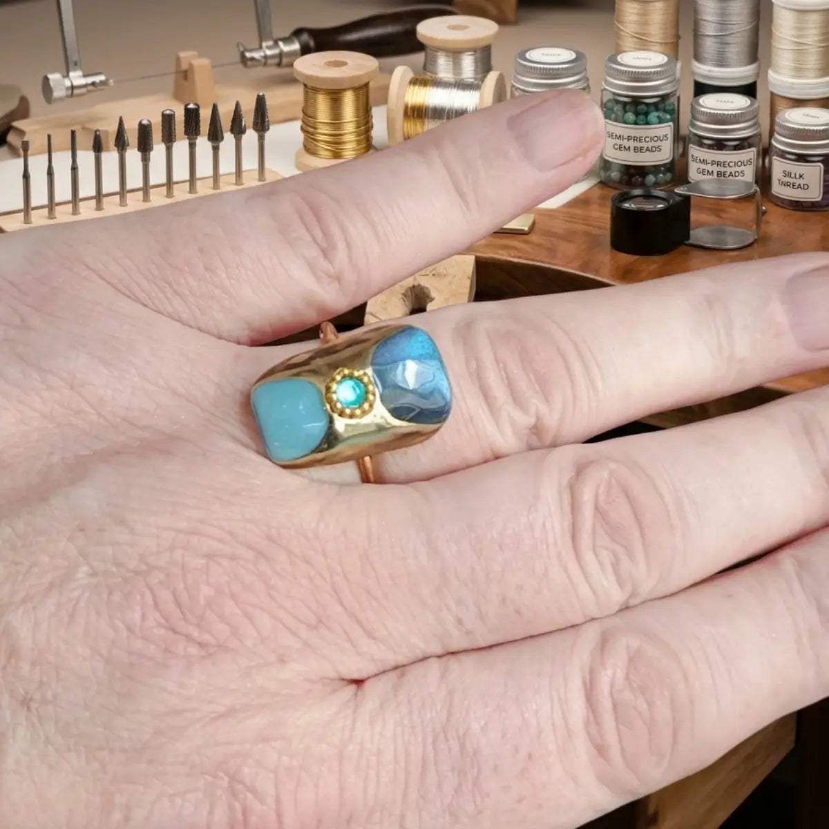 Bague chevalière large dorée avec deux pierres bleues turquoise et détail central vert serti, tenue en main dans un atelier de bijouterie artisanale
