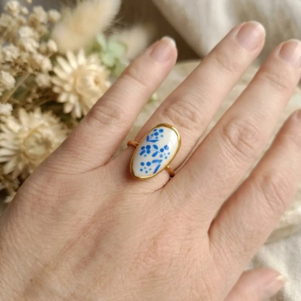 Bague ovale dorée avec médaillon blanc à motifs floraux bleus peints à la main, portée à l'index, style azulejo artisanal