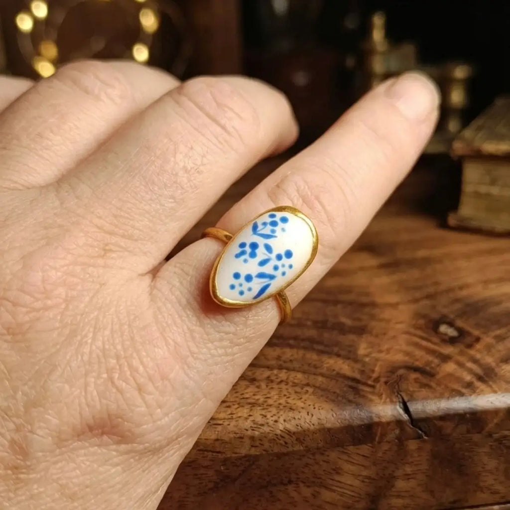 Bague ovale dorée avec médaillon blanc à motifs floraux bleus peints à la main, portée à l'index sur fond bois, bijou artisanal unique