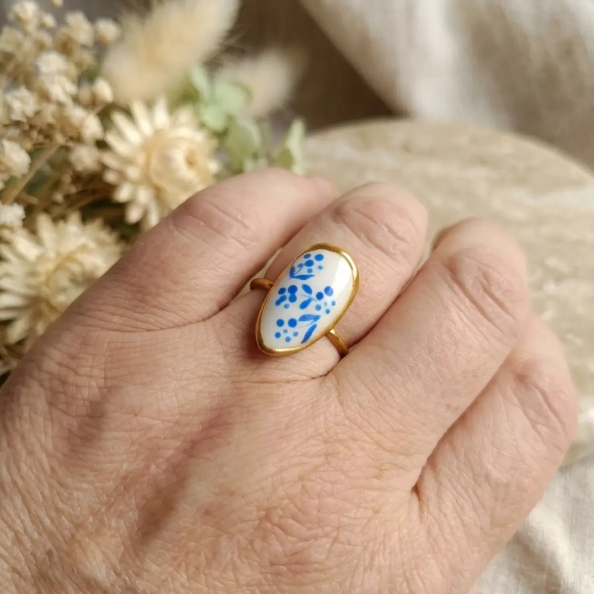 Bague ajustable ovale dorée avec médaillon blanc à motifs floraux bleus peints à la main, vue de côté sur la main, fond fleurs séchées