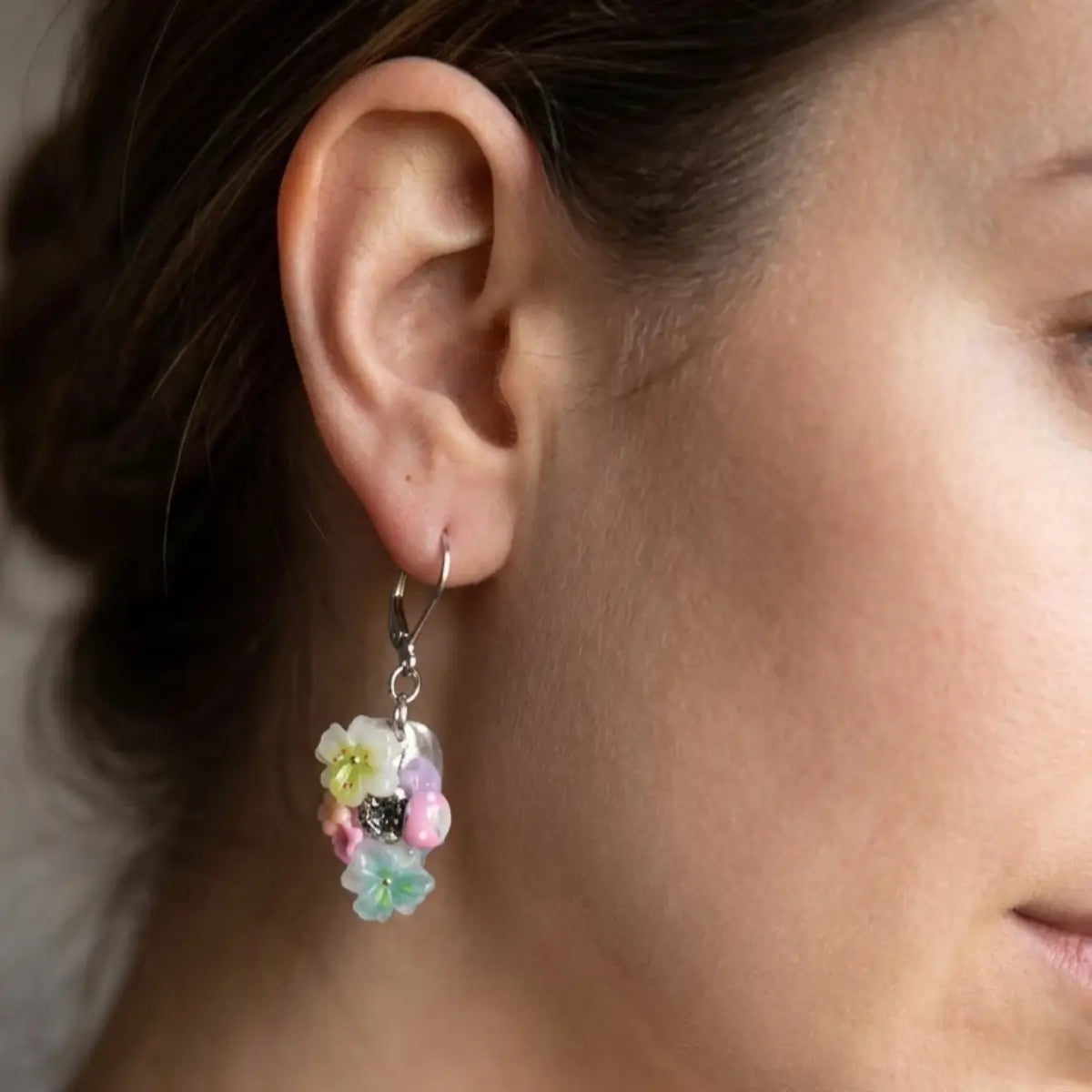 Boucles d'oreilles dormeuses en acier inoxydable argenté portées à l'oreille, avec un pendentif composé de petites fleurs pastel en résine (blanc, jaune, rose, vert menthe) et un strass scintillant au centre.