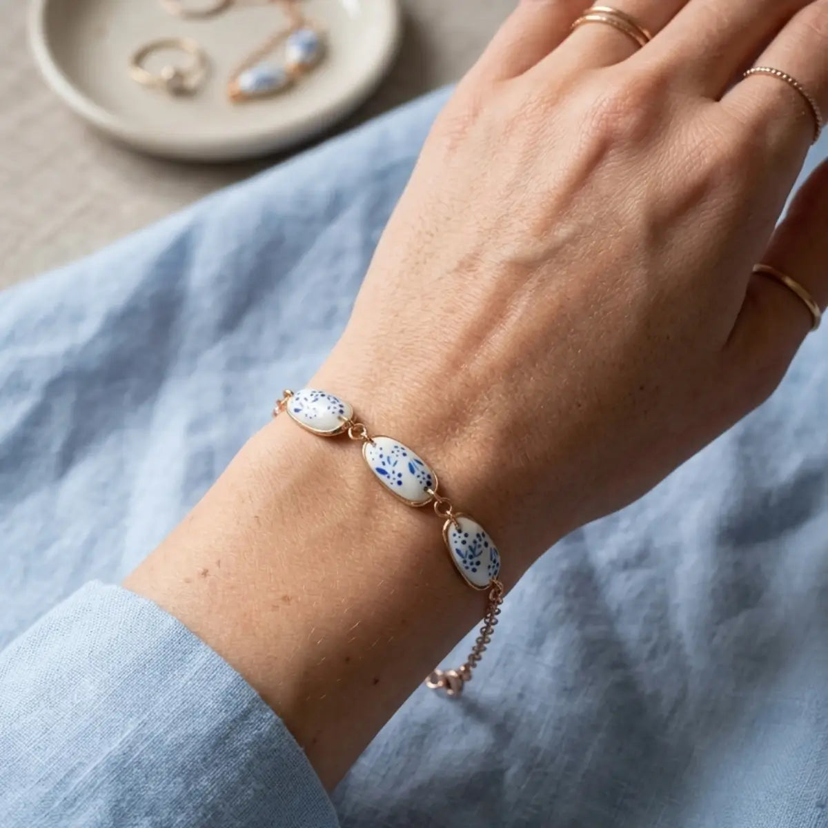 Bracelet chaîne dorée avec trois cabochons amande blancs peints à la main de motifs floraux bleus, porté au poignet sur fond de tissu lin bleu ciel.