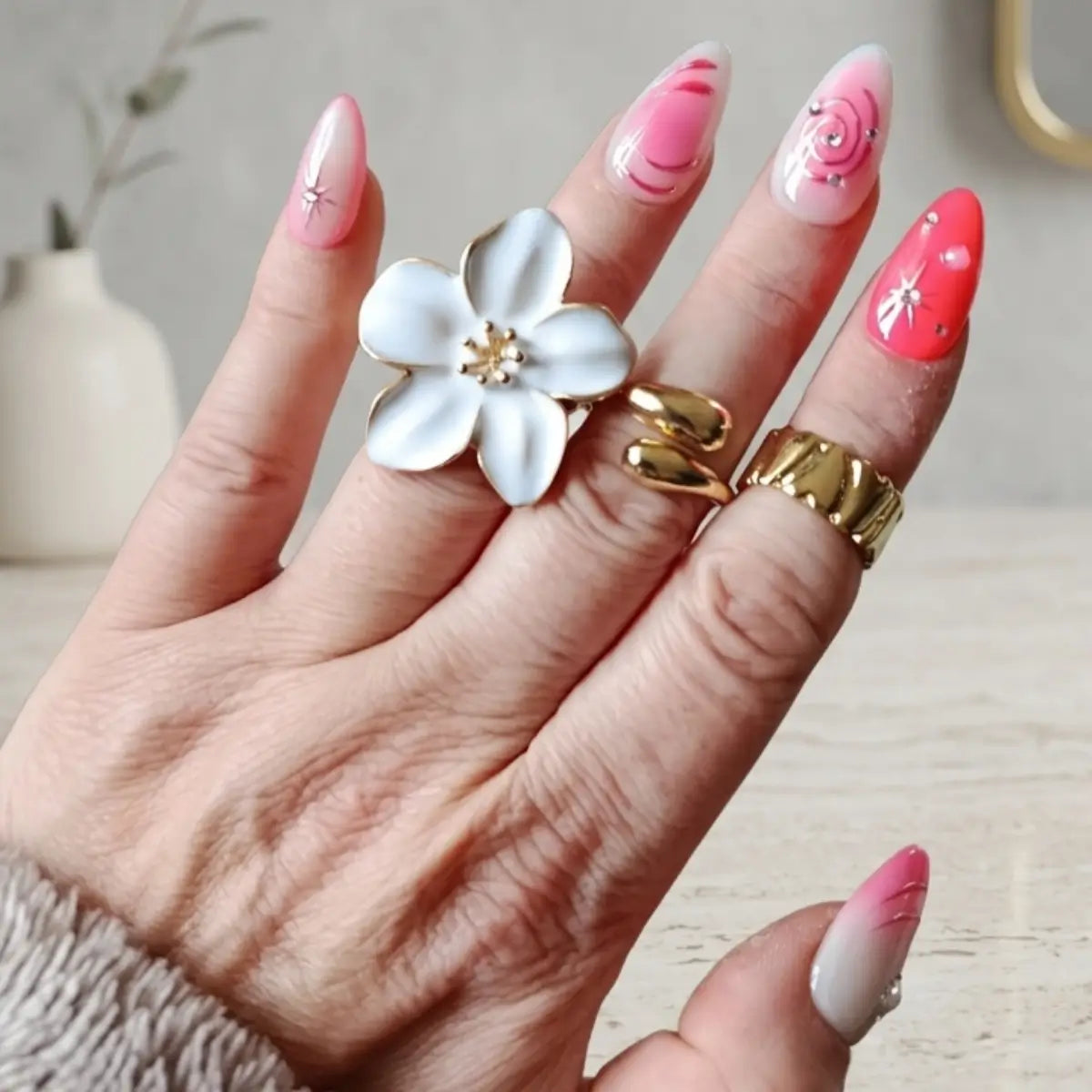 Faux ongles Candy Galaxie Made in France portés sur main mature, mix rose ombré et rouge corail avec spirales et étoiles, bague fleur blanche dorée