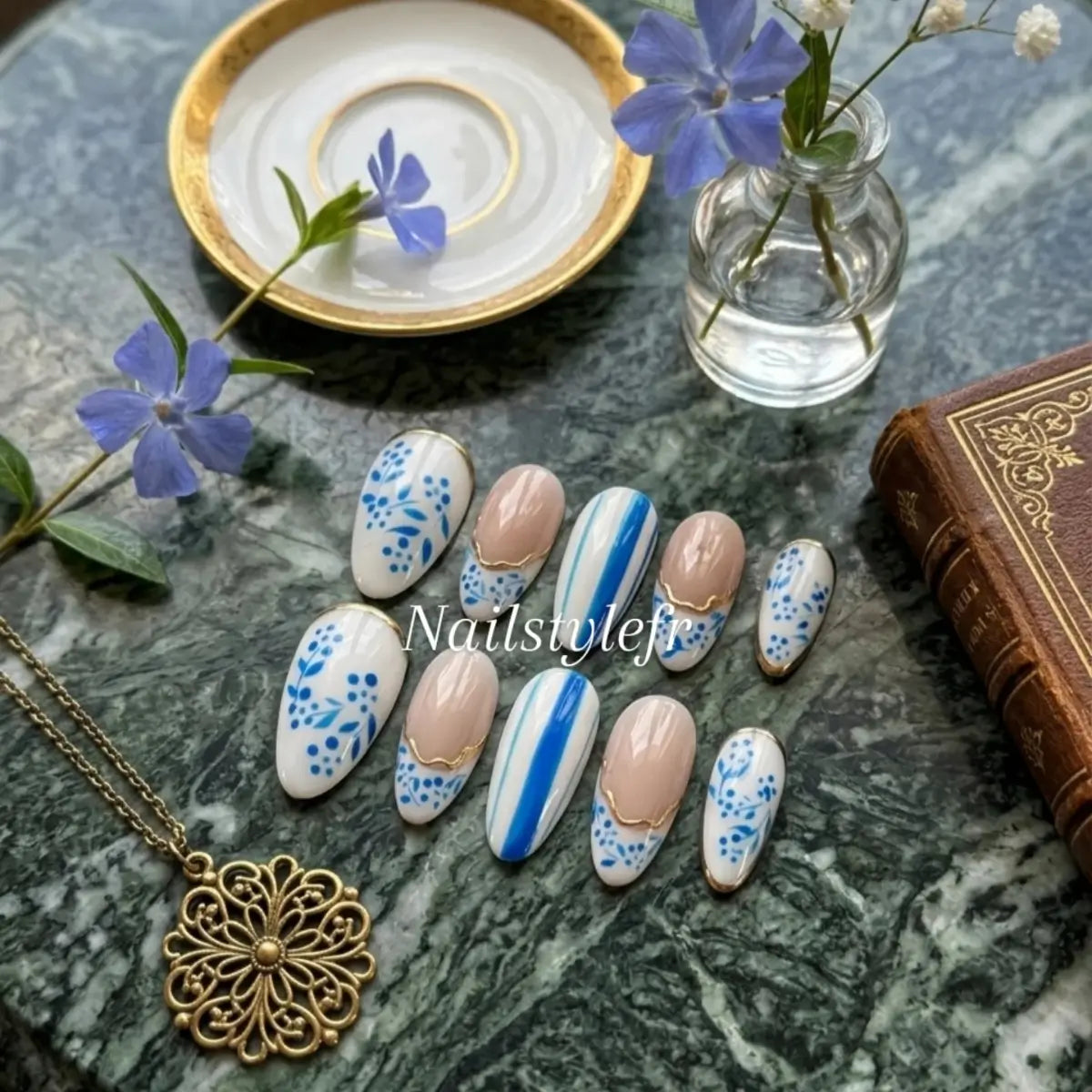 Faux ongles press-on artisanaux motif floral bleu style porcelaine, french nude bordée d'or, présentation sur marbre vert avec carnet ancien et collier vintage