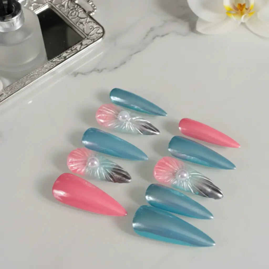 Faux ongles Sinay stiletto bleu et rose - détail des motifs coquillage 3D et strass sur fond marbre blanc.