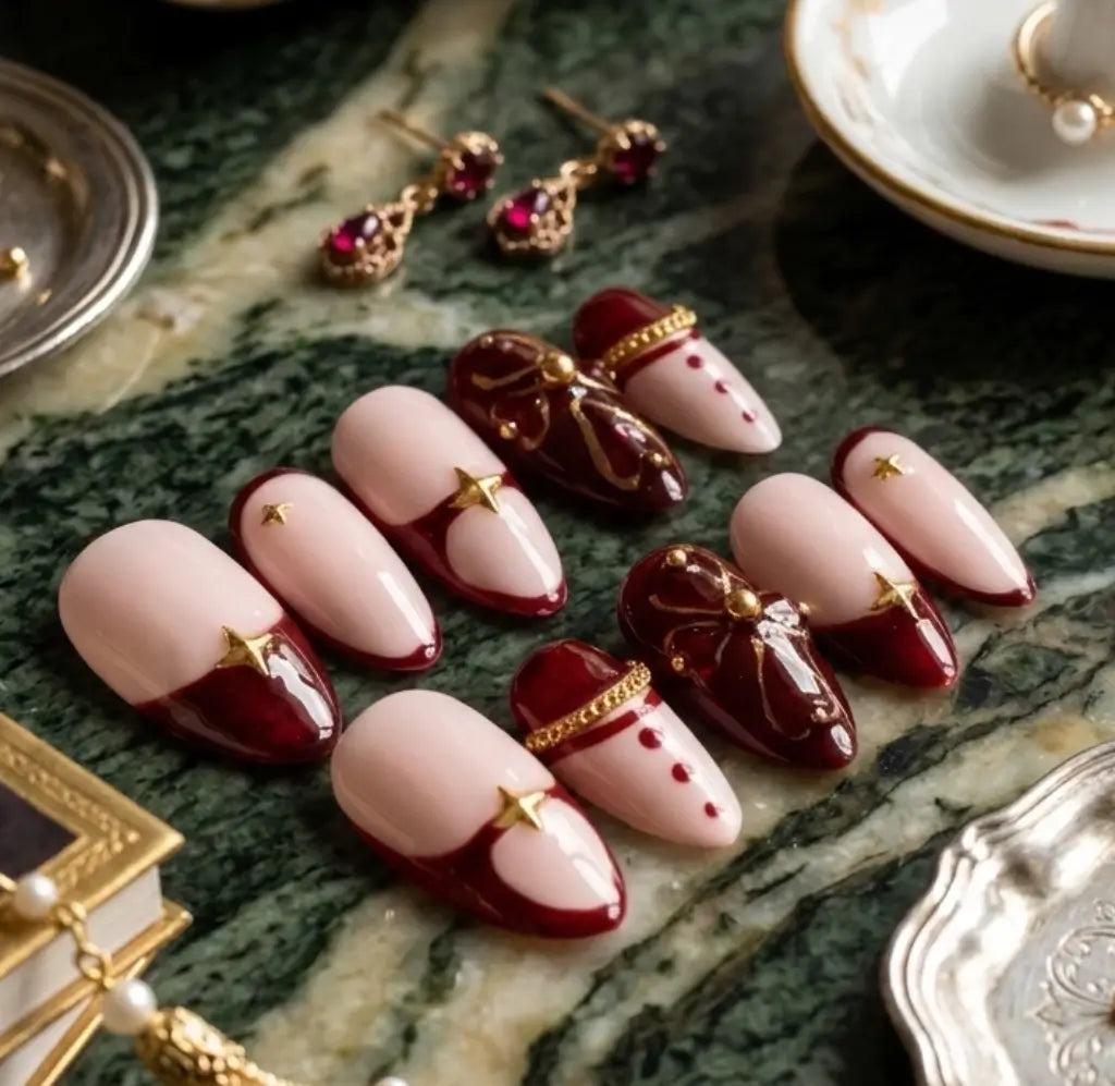 Press-on nails Velvet Rouge – set complet faux ongles bordeaux et nude avec nœud doré et strass, présentation sur marbre vert avec boucles d'oreilles grenat.