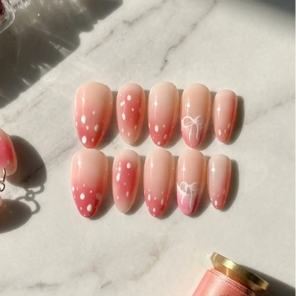 Set de faux ongles forme amande Bambi, dégradé rose pêche avec pois blancs et nœud, posés sur marbre blanc avec bracelet assorti et nœud doré