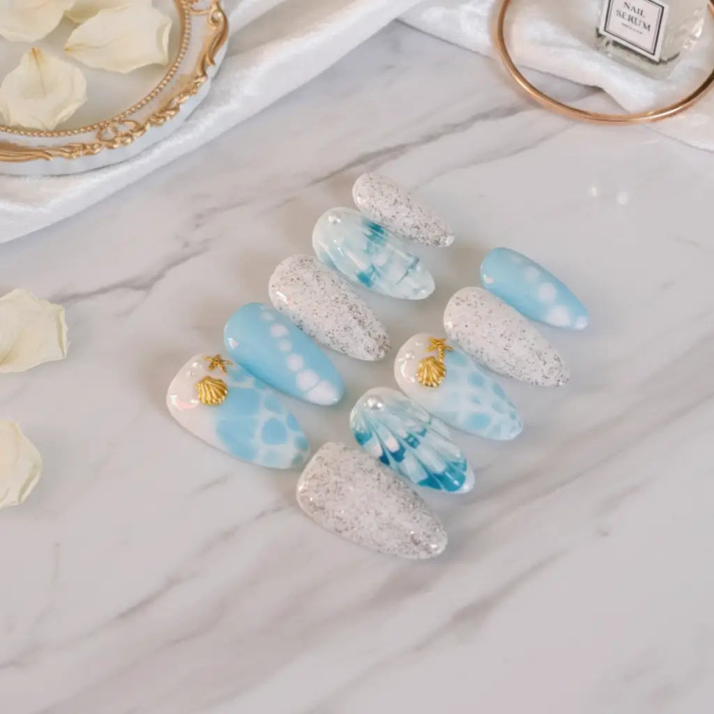 Faux ongles Nérina portés - design aquatique bleu et blanc avec étoiles de mer et coquillages dorés.
