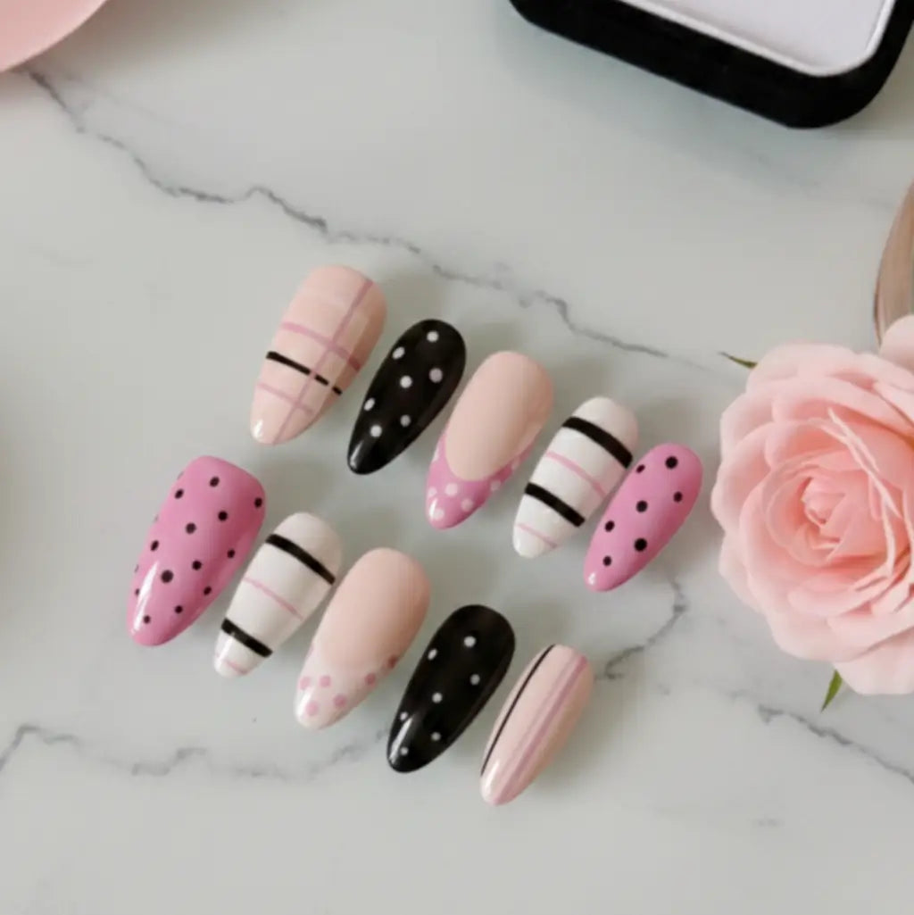 Set de faux ongles réutilisables forme amande avec motifs à pois et rayures, teintes rose poudré, blanc et noir, présentés sur fond marbre avec rose.