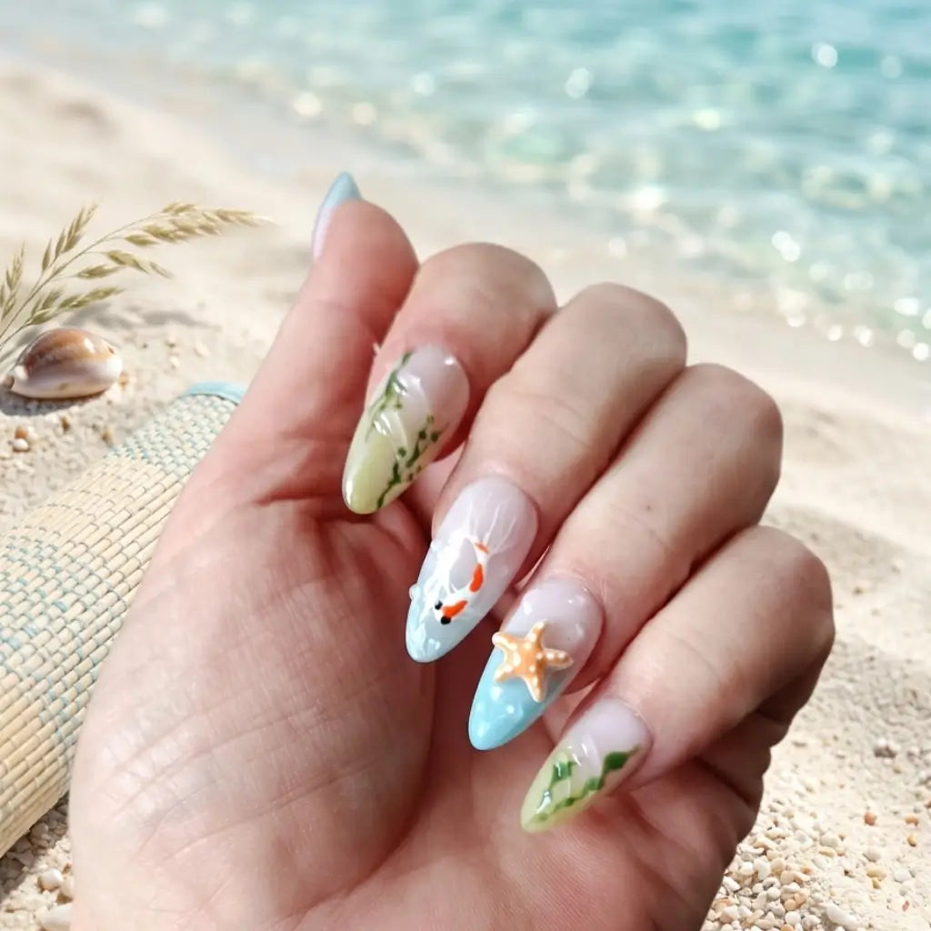 Press-on nails Aqua Flora motif aquatique avec poissons koi, algues et étoile de mer, forme amande longue portés sur plage.
