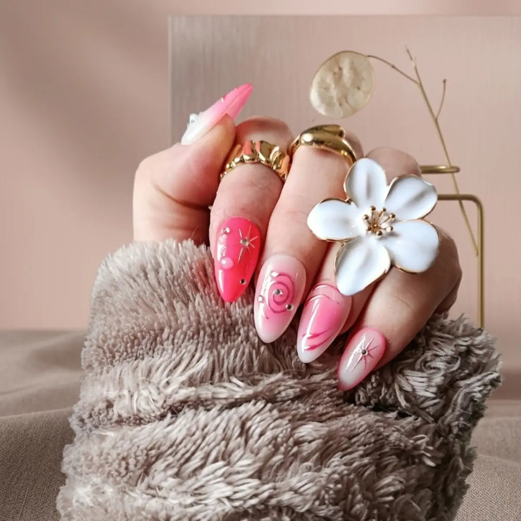 Faux ongles press-on Candy Galaxie en forme amande, dégradé rose et rouge corail avec étoiles dorées et strass, portés avec bagues dorées et fleur blanche.