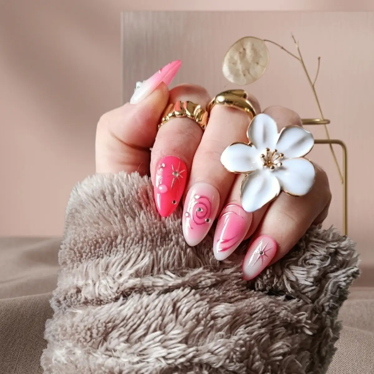 Faux ongles press-on Candy Galaxie en forme amande, dégradé rose et rouge corail avec étoiles dorées et strass, portés avec bagues dorées et fleur blanche.