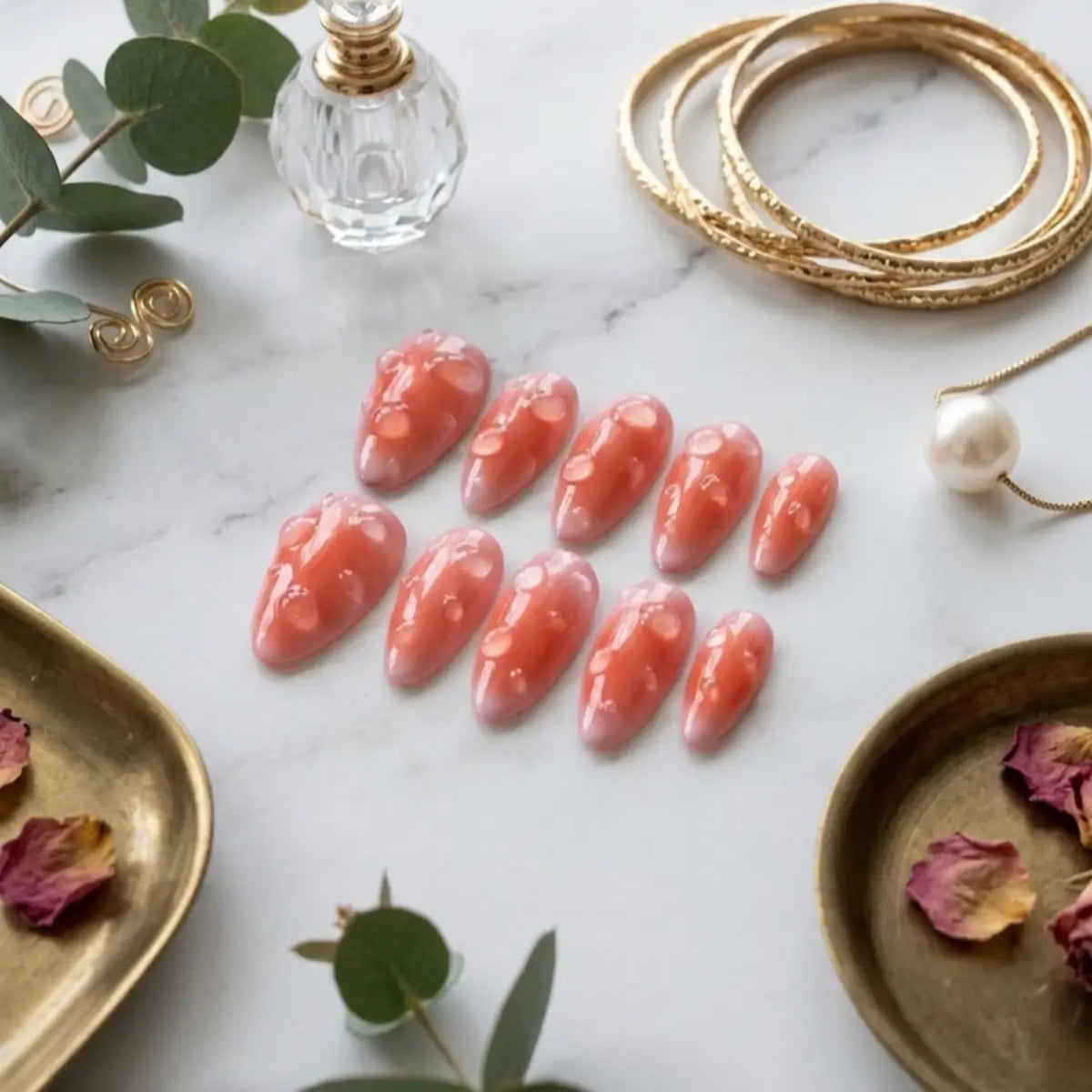 Faux ongles Velvet Peach en flat lay luxe – set corail pêche entouré de pétales de rose, bracelets dorés et perle sur marbre blanc