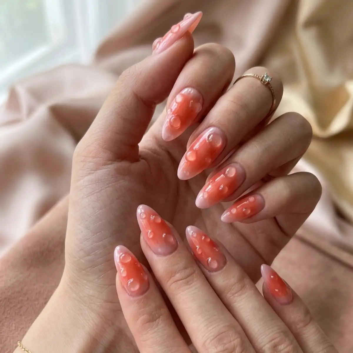 Press-on nails Velvet Peach portés sur deux mains – ongles corail nacré forme amande avec motifs bulles, fond tissu doré