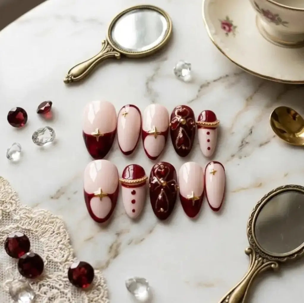 Faux ongles Velvet Rouge forme amande – bordeaux, rose nude et ornements dorés, posés sur marbre avec grenats et miroirs vintage.