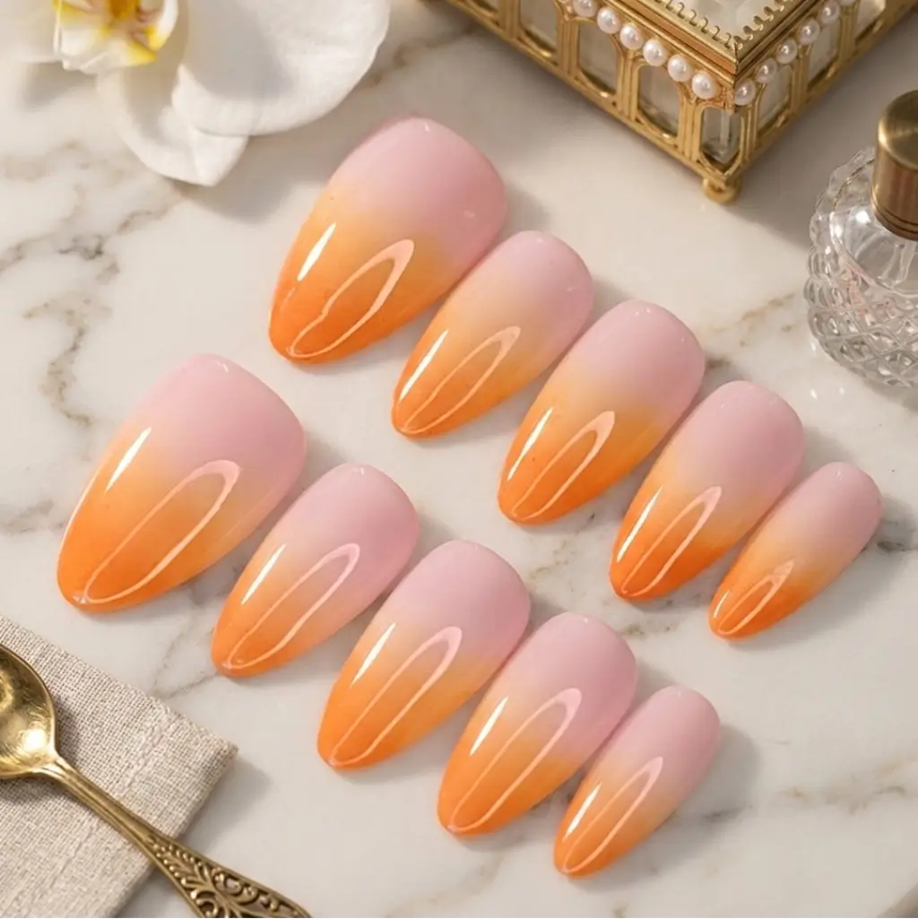 Faux ongles Peach Glow forme amande courte avec dégradé rose poudré vers orange abricot sur marbre blanc avec orchidée.