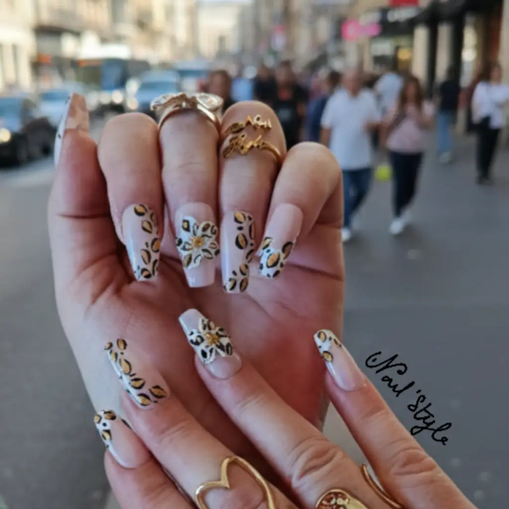 Press-on nails Safari Petals avec motif léopard doré et noir sur base nude rosé, portés en extérieur avec bagues dorées - Nail'Stylefr.