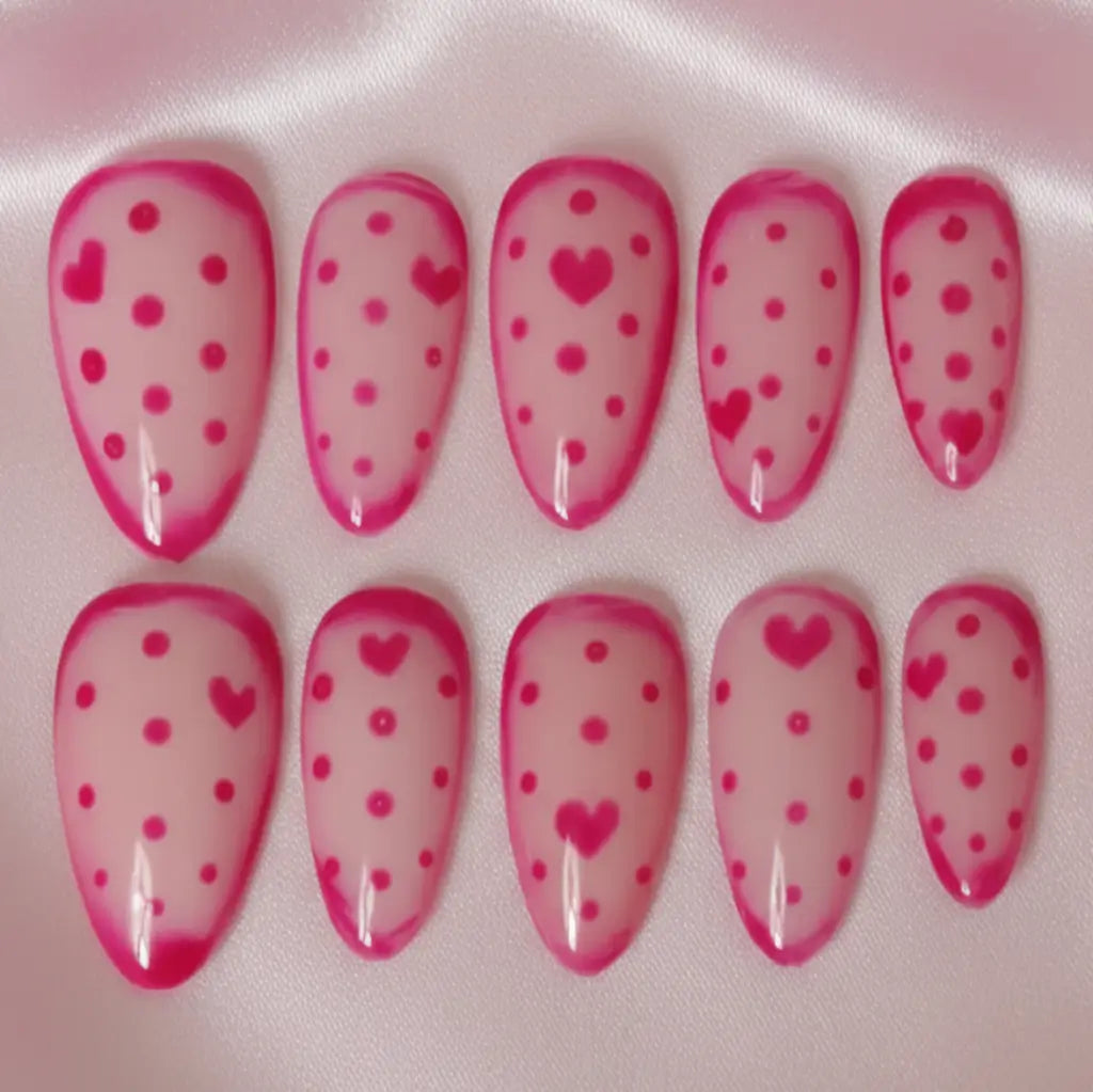 Set complet de 10 faux ongles réutilisables rose avec motifs cœurs et pois sur fond rose transluside. Gros plan sur le design. Forme Amande.