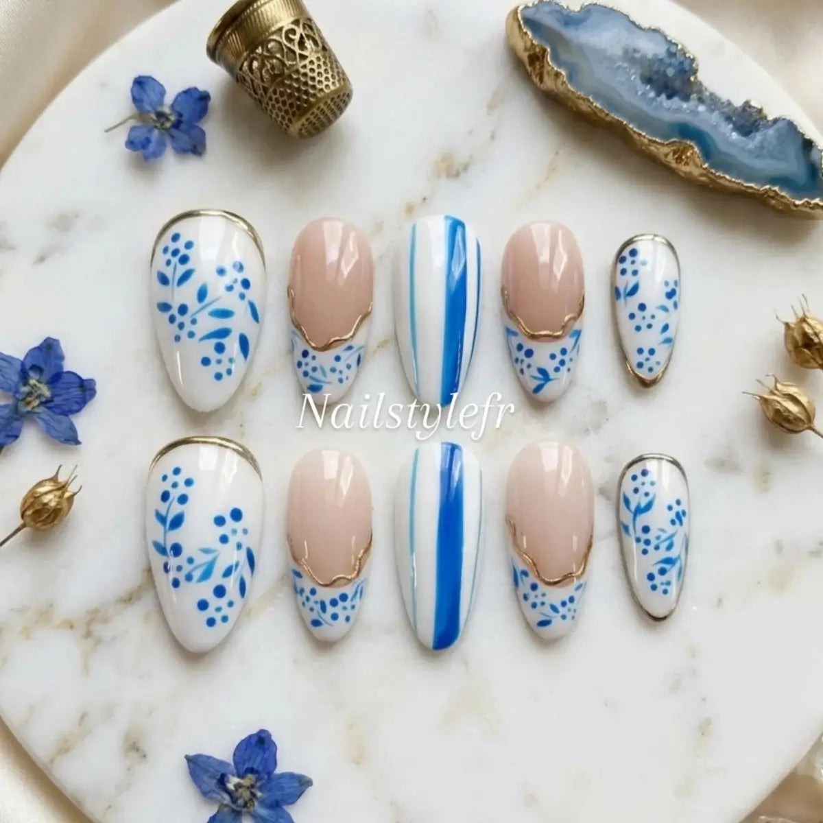 Set de faux ongles press-on forme amande, motif fleurs bleues peintes à la main sur fond blanc, french dorée et accent rayures bleues, posés sur marbre blanc avec fleurs séchées