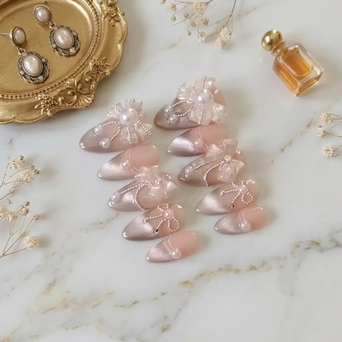 Faux ongles press-on amande cat eye nude rosé avec nœuds en organza et motifs chaînettes nacrées, posés sur marbre