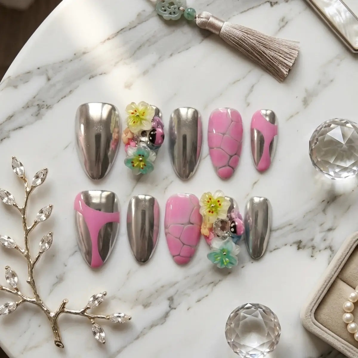 Kit de 10 faux ongles press-on forme amande posés sur marbre blanc : finition chrome argenté miroir, rose craquelé et ongles accent avec fleurs pastel 3D (jaune, rose, vert) et coccinelle en résine