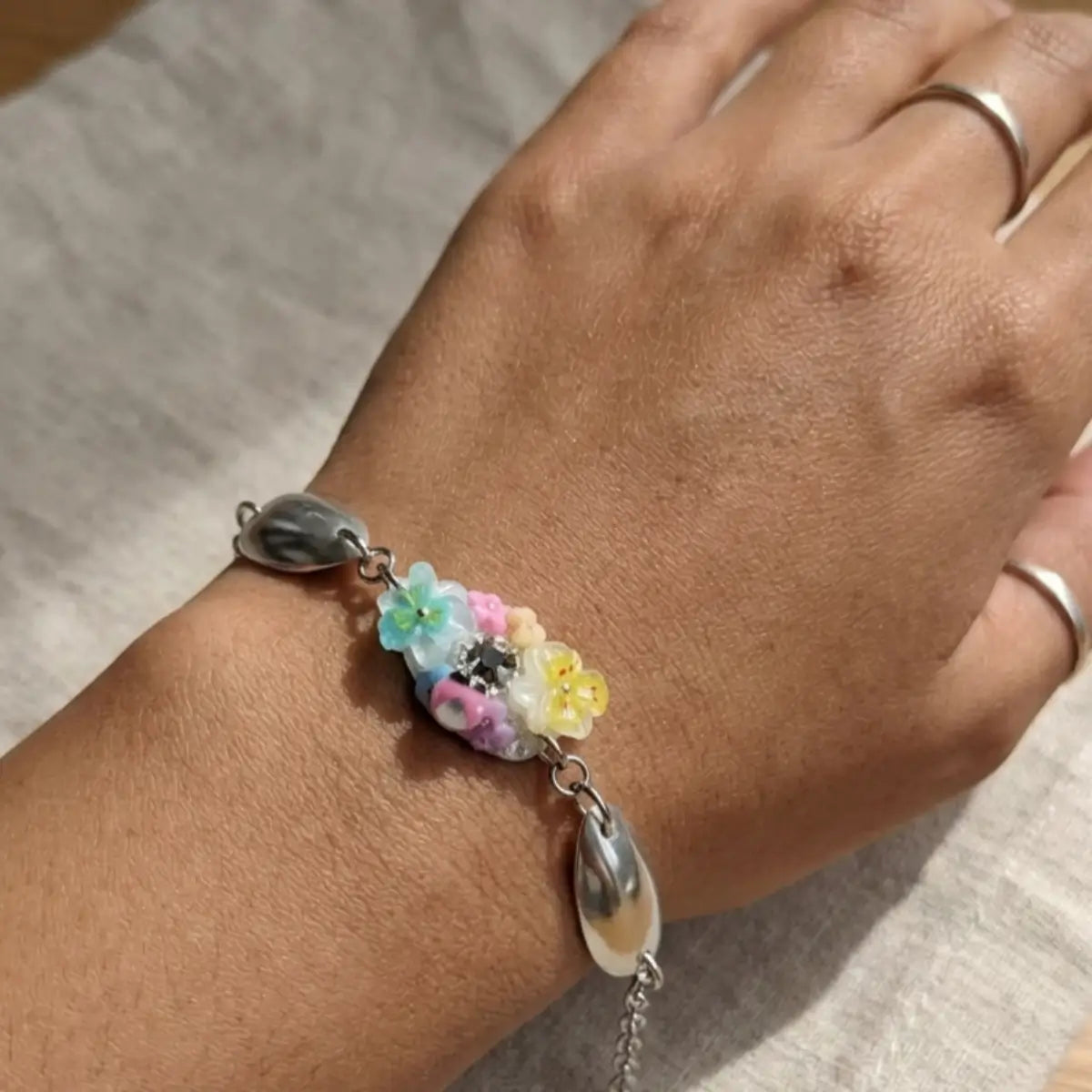 Bracelet en acier inoxydable argenté porté au poignet, avec un cabochon central orné de fleurs pastel multicolores (bleu, rose, jaune, blanc) et un strass, encadré de deux perles ovales en nacre grise.