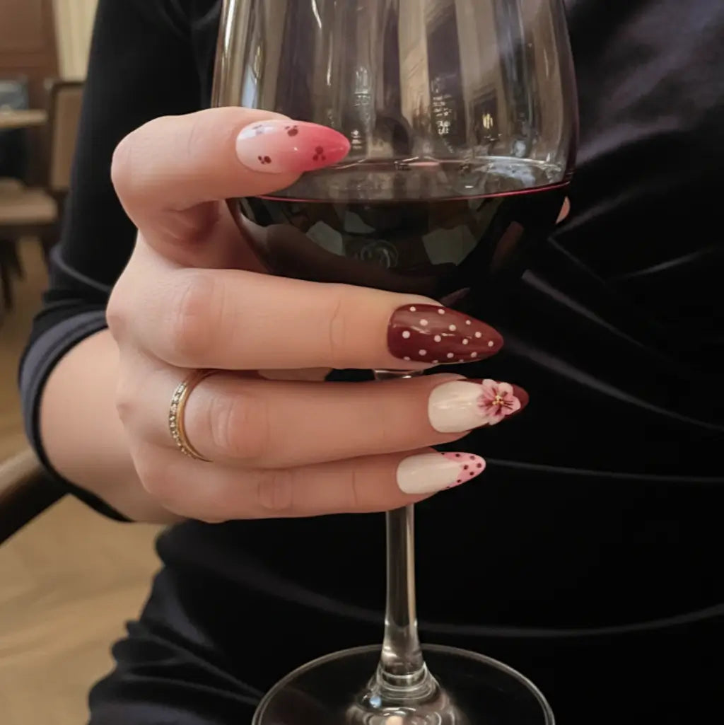 Faux ongles pressés réutilisables bordeaux et rose poudré avec motifs pois et fleurs, portés avec un verre de vin. Made in France.