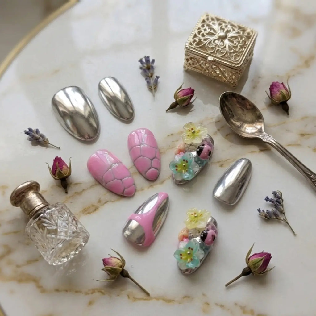 Faux ongles press-on forme amande disposés sur plateau marbré entourés de boutons de roses et lavande séchés : chrome argenté, rose craquelé et décors floraux pastel 3D avec coccinelle.